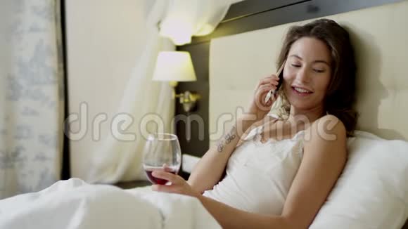 漂亮的女人躺在床上一边喝着红酒一边用智能手机说话视频的预览图