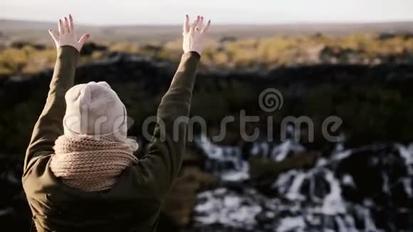快乐的女人站在冰岛的Barnafoss瀑布附近跳着年轻女性摘下帽子举手视频的预览图