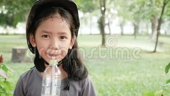 可爱的亚洲小女孩戴着粉红色的头盔在自然公园喝着幸福的水视频的预览图