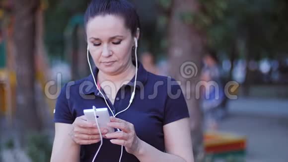 一个戴着耳机的女人站在公园里把手指滚动在智能手机屏幕上视频的预览图