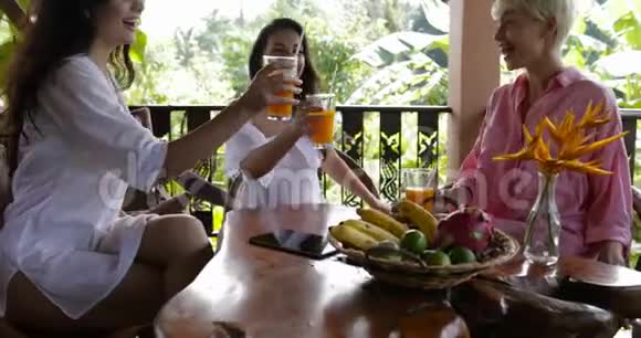 在热带森林的夏季露台上年轻女孩会在早餐时一起品尝带果汁的玻璃杯美女们视频的预览图