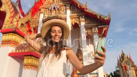 穿着白裙戴着大草帽的年轻混血旅游女孩在泰国用手机拍照视频的预览图