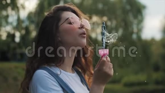 美丽的年轻女子戴着太阳镜在户外吹肥皂泡到处旋转近景慢动作视频的预览图