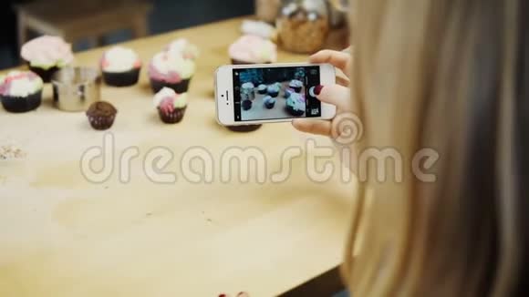近距离观看女性与她烘焙的纸杯蛋糕拍照年轻女子在智能手机上使用相机视频的预览图