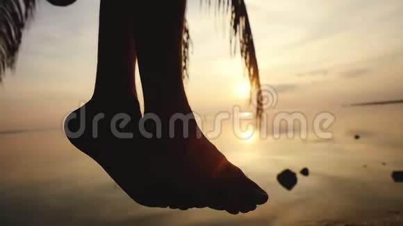 接近男性腿的轮廓在美丽的夕阳下年轻人坐在棕榈树上在热带海滩挥舞着双腿视频的预览图