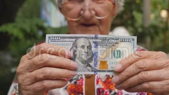 戴着眼镜的老妇人把100美元的钞票放进相机在户外微笑奶奶在外面拿着现金视频的预览图