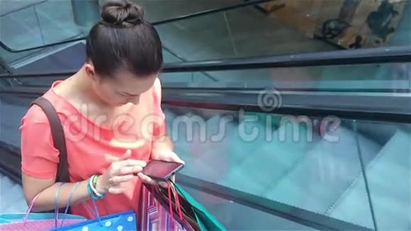 年轻女子在购物中心的自动扶梯上使用智能手机并使用智能手机城市生活方式保持联系视频的预览图