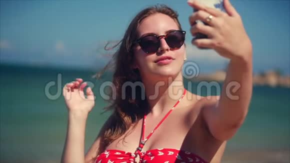 年轻美丽的布鲁内特女士站在沙滩上夏天看着她的手机和自拍背景蓝色视频的预览图