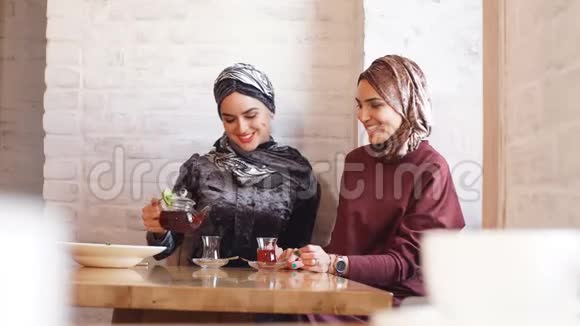 两个漂亮的穆斯林女人在咖啡馆里戴着头巾坐在桌子旁的沙发上聊天视频的预览图