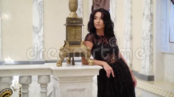 年轻的黑发女子穿着黑色晚礼服在宫殿或酒店的大理石石梯前摆姿势视频的预览图
