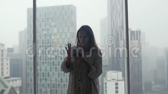 雨中美丽的年轻女子站在窗边在城市的背景下使用智能手机3840x2160视频的预览图