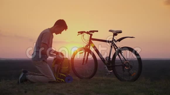 一个年轻人把东西放在背包里日落时骑自行车坐下视频的预览图