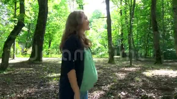 微笑的孕妇走在公园里嫩嫩的大肚子在树上晒太阳视频的预览图