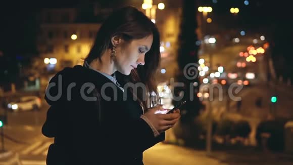 年轻的美女晚上站在市中心的交通道路的背景下使用智能手机视频的预览图
