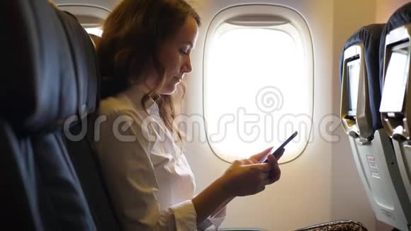 在飞机上使用智能手机的旅游女孩坐在飞机的窗户附近视频的预览图