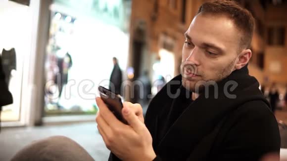 年轻的帅哥留着胡子坐在咖啡馆里在市中心度过闲暇时光使用智能手机视频的预览图