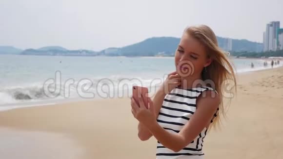 年轻漂亮的苗条女人一头金发穿着黑白连衣裙在海边的手机上自拍视频的预览图