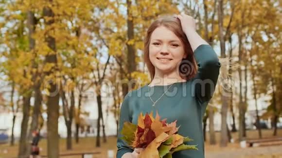 年轻漂亮的女孩穿着礼服带着一束秋叶在镜头中摆姿势在城市公园欣赏秋天视频的预览图