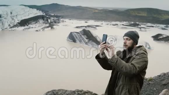年轻英俊的男子徒步旅行在智能手机上拍摄了冰岛的Vatnajokull冰泻湖和冰川视频的预览图