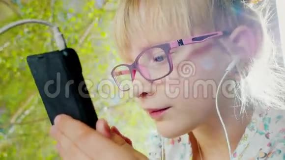 一个戴着粉红色眼镜的积极开朗的女孩的肖像她在阳光下用窗户上的电话春天的心情视频的预览图