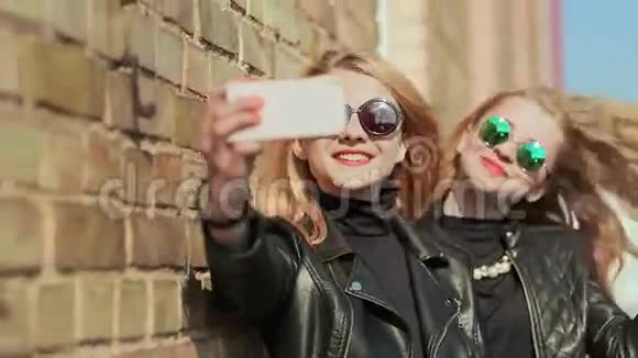 两个戴着太阳镜和时髦皮夹克的女朋友从靠近砖墙的手机上拍照视频的预览图