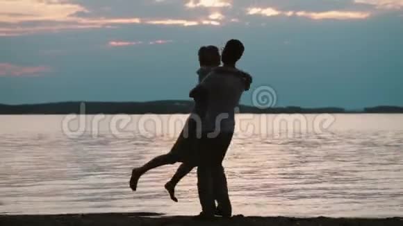 年轻的幸福夫妇在海滩上的夕阳下拥抱迷人的男人和女人在傍晚的河岸上旋转视频的预览图