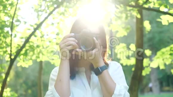 一位身穿白衬衫笑容可掬的女摄影师正在公园里柔和的背景下拍摄照片视频的预览图