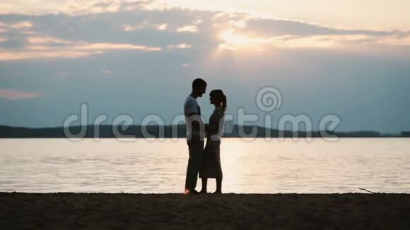 晚上一对年轻幸福的夫妇拥抱在河岸上的剪影男人和女人花时间在海滩上视频的预览图