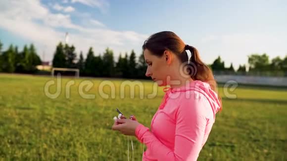 三合一视频日落时分戴着耳机和智能手机的女人穿过体育场视频的预览图
