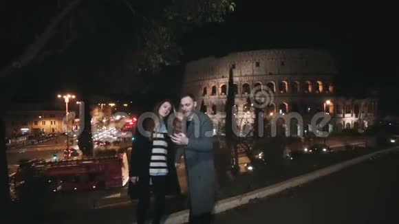 年轻漂亮的情侣站在意大利罗马的斗兽场附近晚上在智能手机上谈论自拍照片视频的预览图