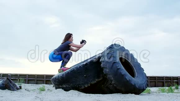 运动的女人穿着T恤蓝色的腿用一个大拖拉机轮子做力量练习她在跳关于视频的预览图