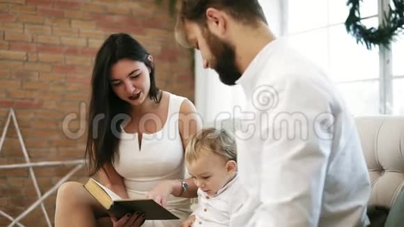年轻的母亲为儿子和丈夫读书一家人坐在圣诞树旁看圣诞故事快乐视频的预览图