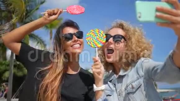 两个年轻的少女在热带海滩用手机和舔棒棒糖做自拍高清慢速运动苏视频的预览图