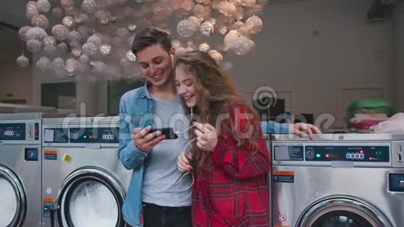 一对美丽的情侣在洗衣间听着电话里的音乐看着视频故事英俊的年轻人视频的预览图