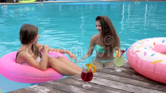 带着鸡尾酒的快乐女朋友在泳池边亲吻空气夏天在游泳池里休息穿泳衣的朋友视频的预览图