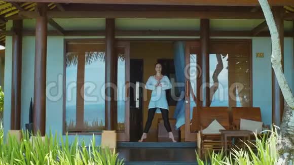 女人正在练习瑜伽姿势伸展在巴厘岛海滩上的平房门廊上大自然的声音视频的预览图