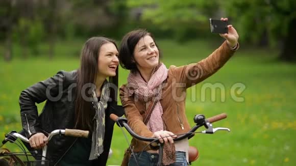 两个穿着夹克和围巾的女自行车手正在用智能手机拍照带着自行车的年轻漂亮的布鲁纳视频的预览图