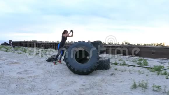运动的女人穿着T恤蓝色的腿用一个大拖拉机轮子做力量练习她在跳关于视频的预览图