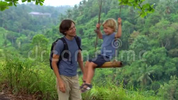 一个年轻人在热带公园的秋千上摆动他的小儿子的慢镜头儿童概念视频的预览图