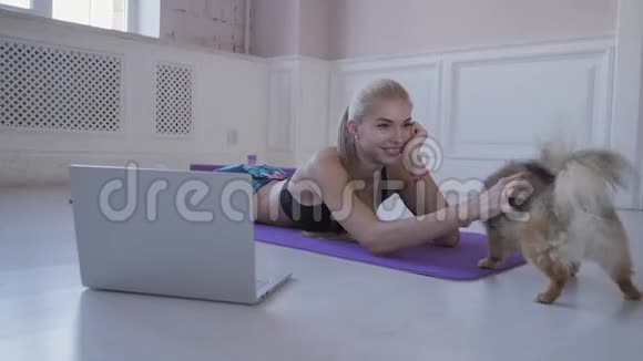 家庭健身年轻女子在运动和玩小狗斯皮兹在网上观看笔记本电脑上的视频视频的预览图