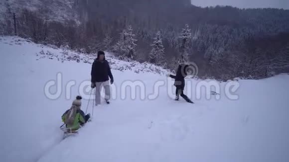 年轻的幸福家庭在雪林里玩得很开心父母把他们的小儿子卷在雪橇上晚上散步视频的预览图