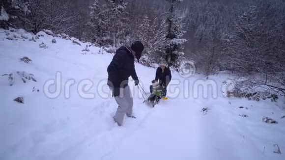 年轻的幸福家庭在雪林里玩得很开心父母把他们的小儿子卷在雪橇上晚上散步视频的预览图