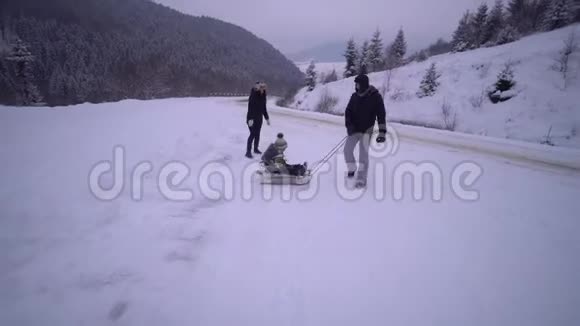 年轻的幸福家庭在雪林里玩得很开心父母把他们的小儿子卷在雪橇上晚上散步视频的预览图