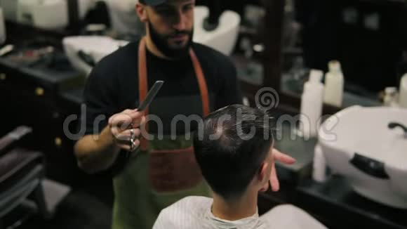 理发店里坐在椅子上理发的一位男客户身穿围裙梳头的胡子理发师时尚视频的预览图