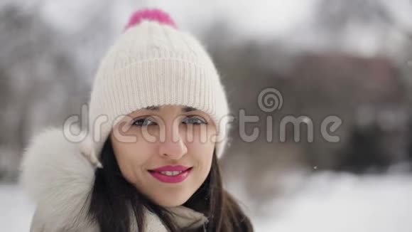 特写黑发年轻女性肖像粉红色嘴唇米色羽绒服帽子在雪地中央公园微笑视频的预览图