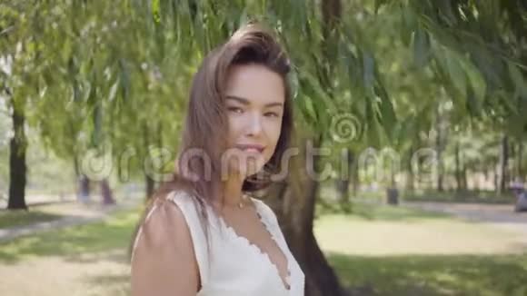 肖像可爱的年轻女孩一头长长的黑发穿着一件白色的夏装站在树枝下视频的预览图