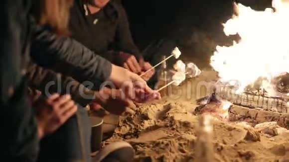 一群多民族的年轻人深夜坐在海边的火堆旁用棍子烤棉花糖视频的预览图