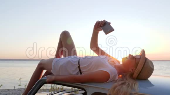 快乐之旅年轻女子躺在屋顶汽车上用电话拍照女性在手机上用复古拍照视频的预览图