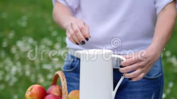 在一片洋甘菊草坪的背景下女人的手把茶从一个白色的罐子里倒入一个白色的杯子一个热水瓶关于视频的预览图