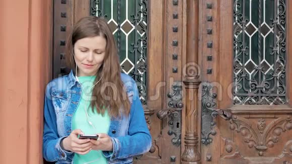 穿着棉夹克的漂亮女孩用智能手机带耳机背景中的老式建筑视频的预览图